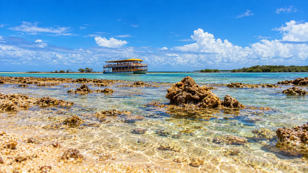 Banco de areia coralínea e piscinas naturais na maré baixa no Parque Marinho de Coroa Alta, em Santa Cruz Cabrália, com escuna ao fundo.